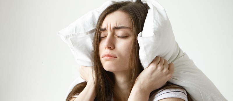 Pretty student girl in striped t-shirt trying to have some sleep, sitting in bedroom with pillow on her head, closing eyes, having tired painful look, irritated with annoying sound. Negative emotions
